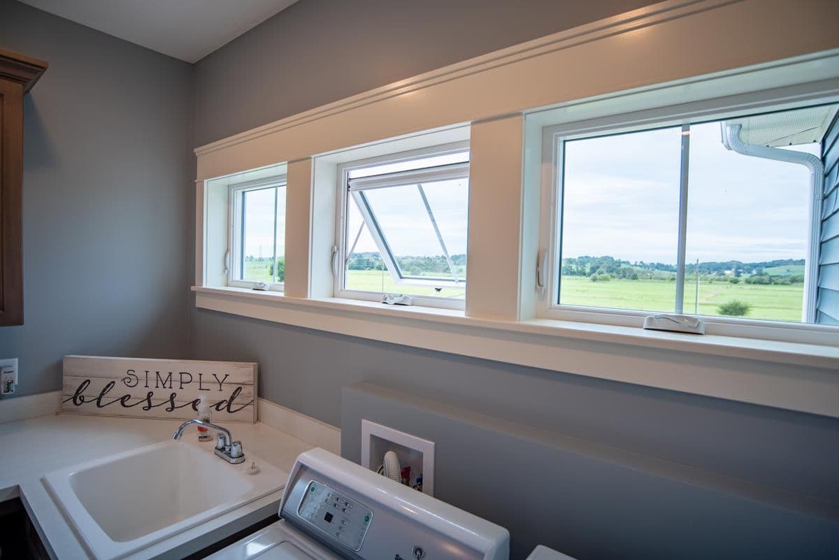 Bathroom with large windows overlooking green fields, white sink and washing machine, wooden sign reading "simply blessed" on counter