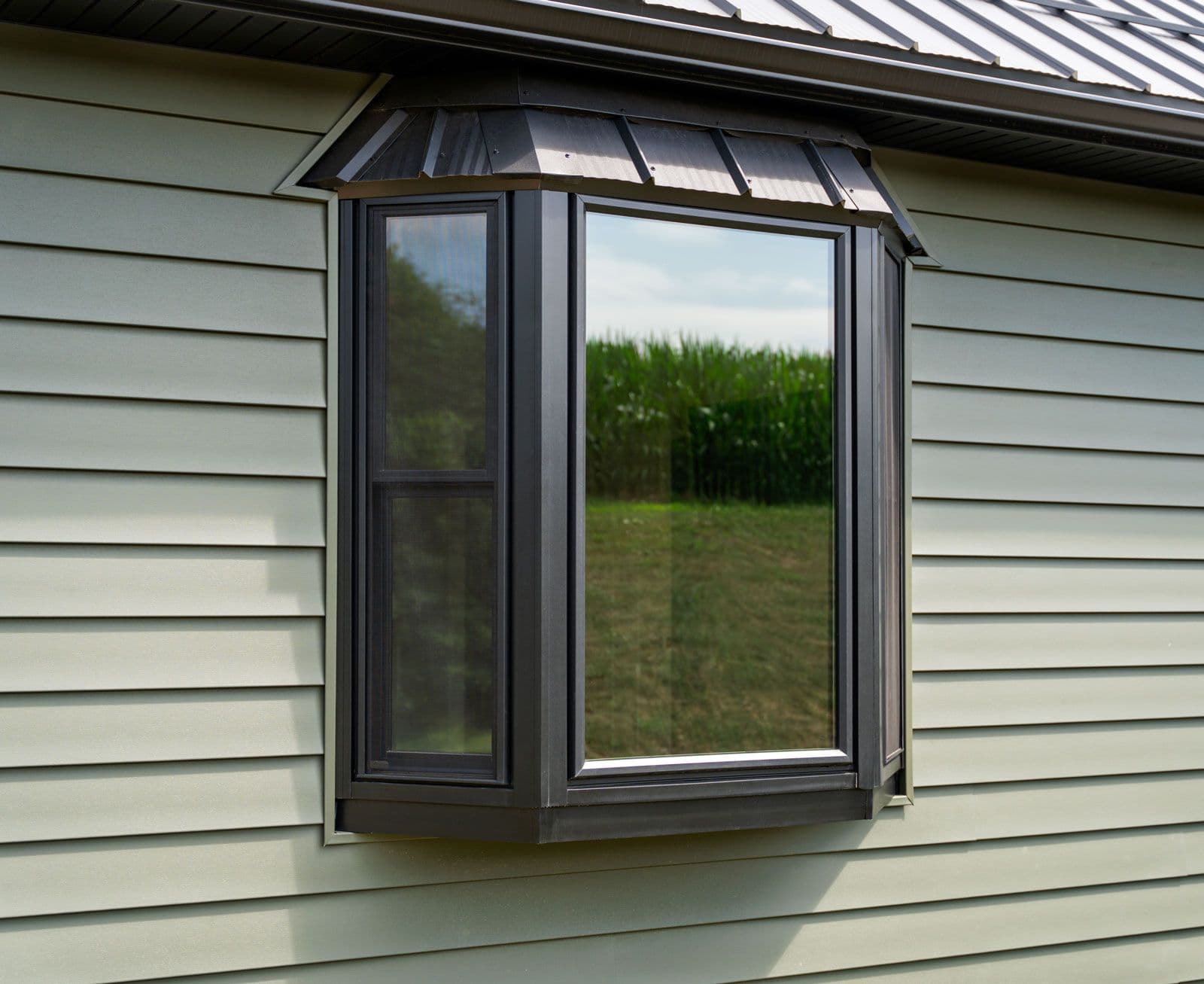 Modern bay window with dark frame and metal awning installed on light beige siding, overlooking rural landscape with field and trees