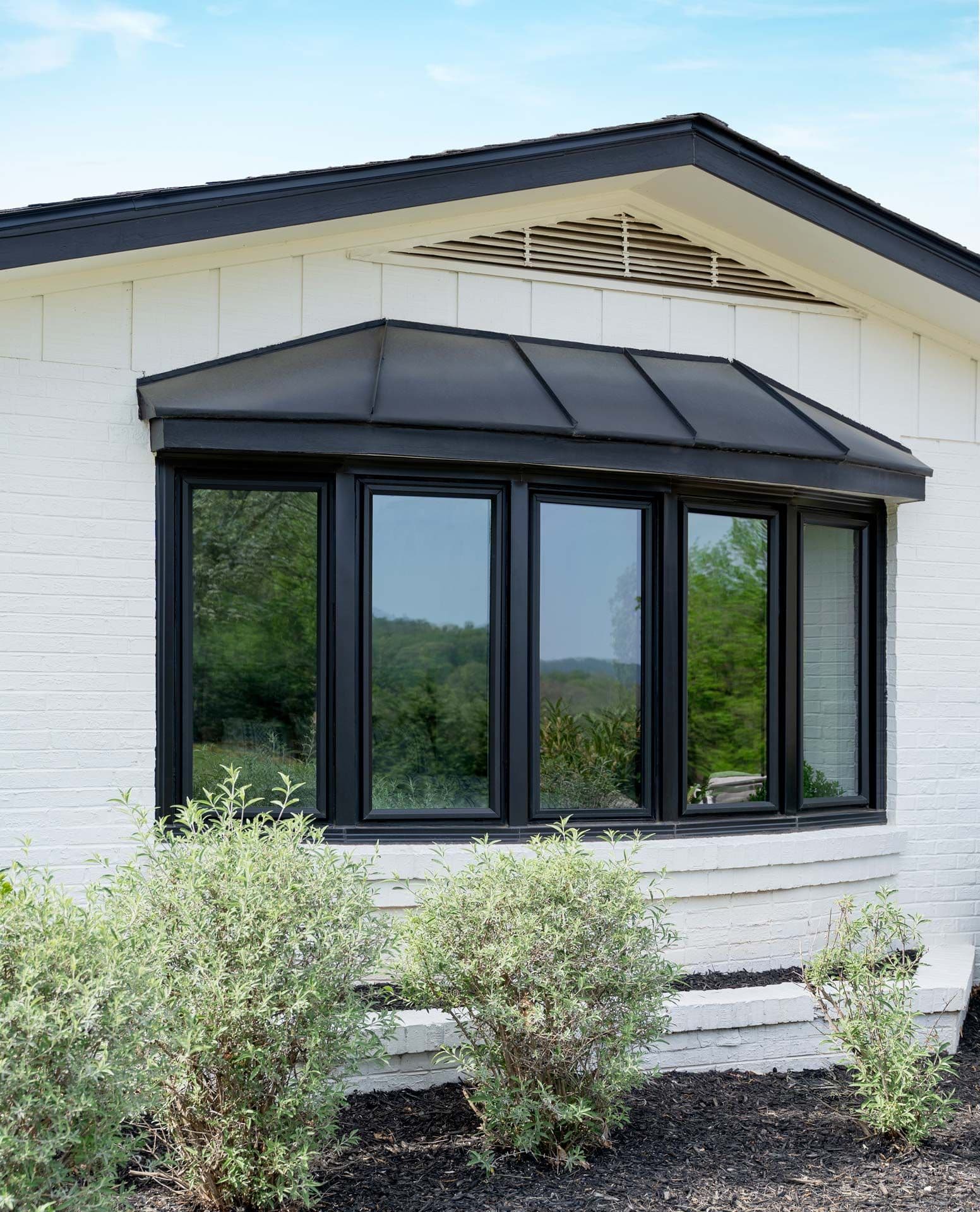 Modern house extension with five black-framed glass doors and a dark metal awning, white brick exterior, and landscaping with shrubs
