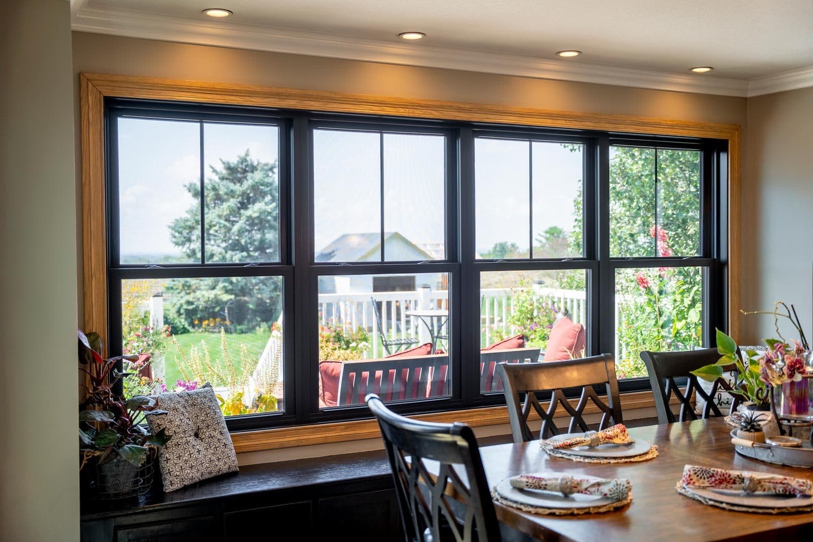 Dining room with large black-framed windows overlooking a backyard garden and patio with seating, wooden window seat with cushion and plants in foreground, dining table set with place settings in immediate foreground
