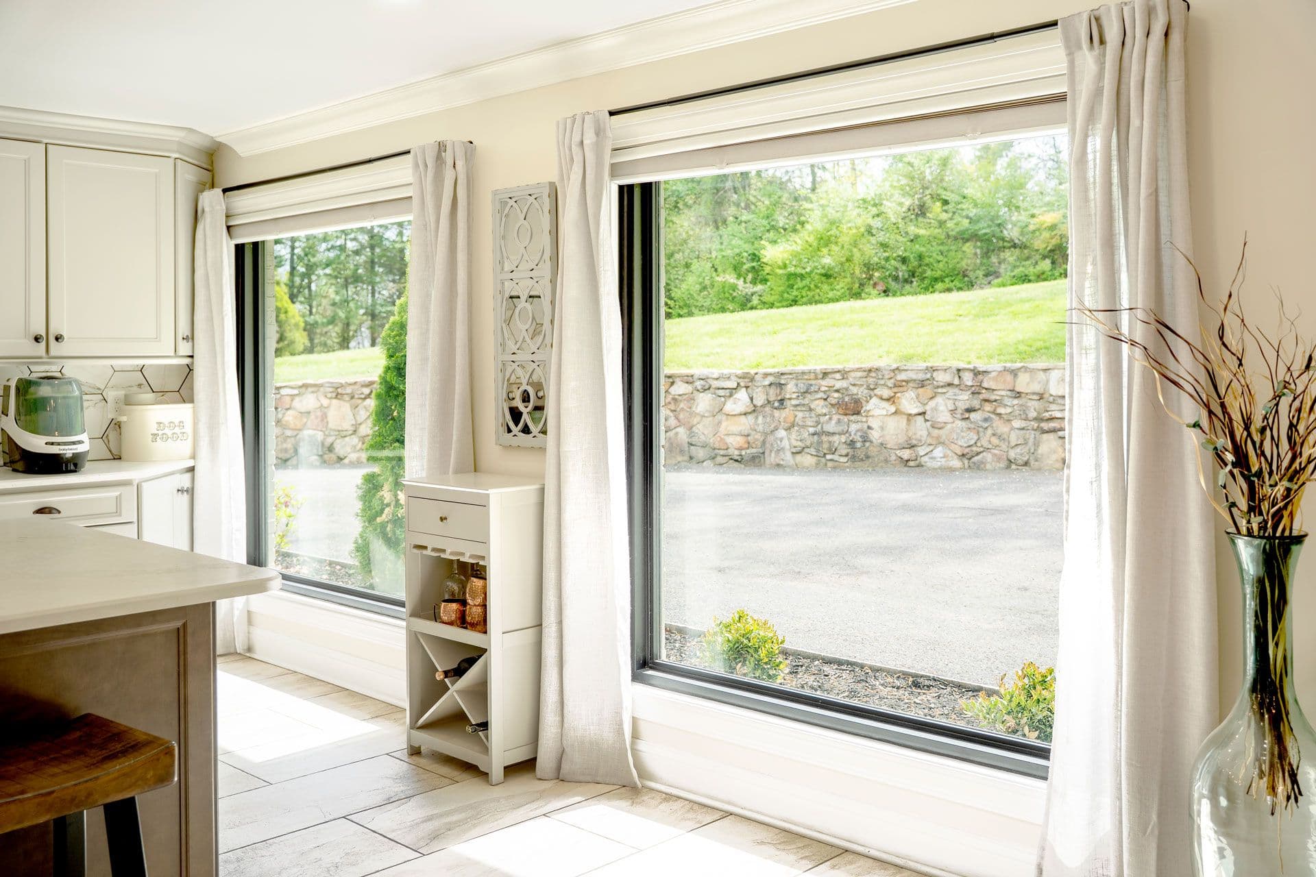 Kitchen with cream cabinetry and cream curtains, featuring large sliding glass doors opening to a patio with stone wall and green landscape beyond