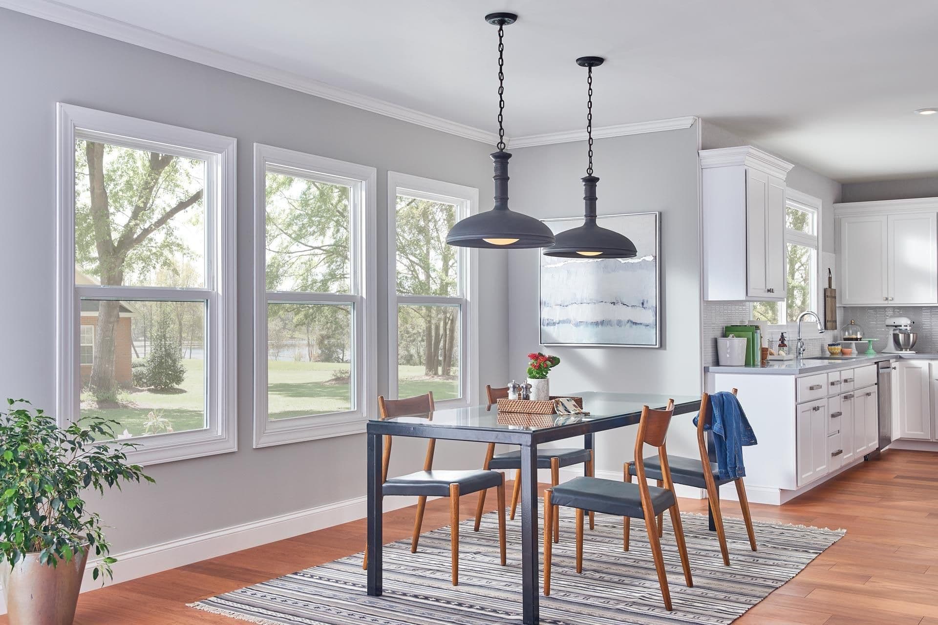 Modern kitchen-dining area with black pendant lights over dining table, wood chairs, large windows overlooking trees, and white cabinetry visible in background